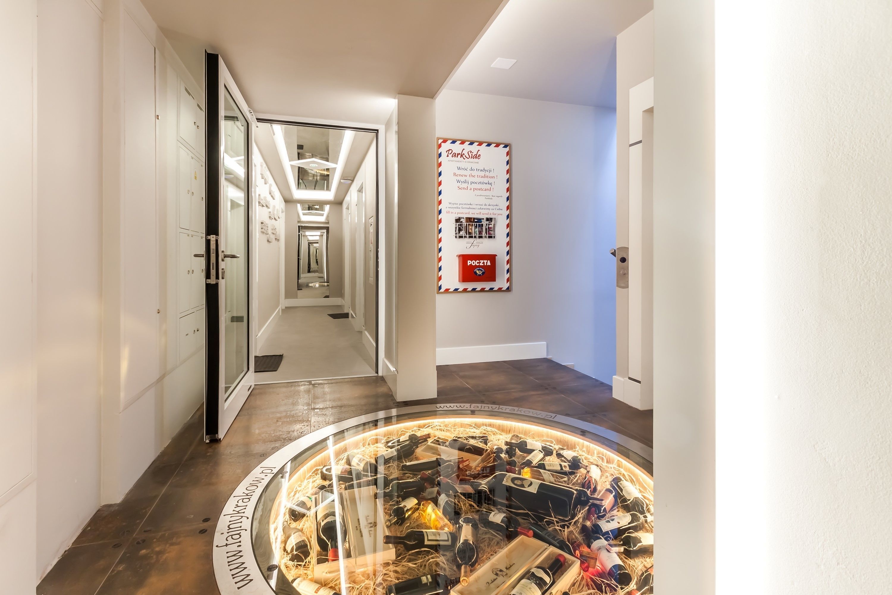 A hotel hallway with a circular display of wine bottles, a glass door leading to a room, and a poster on the wall.