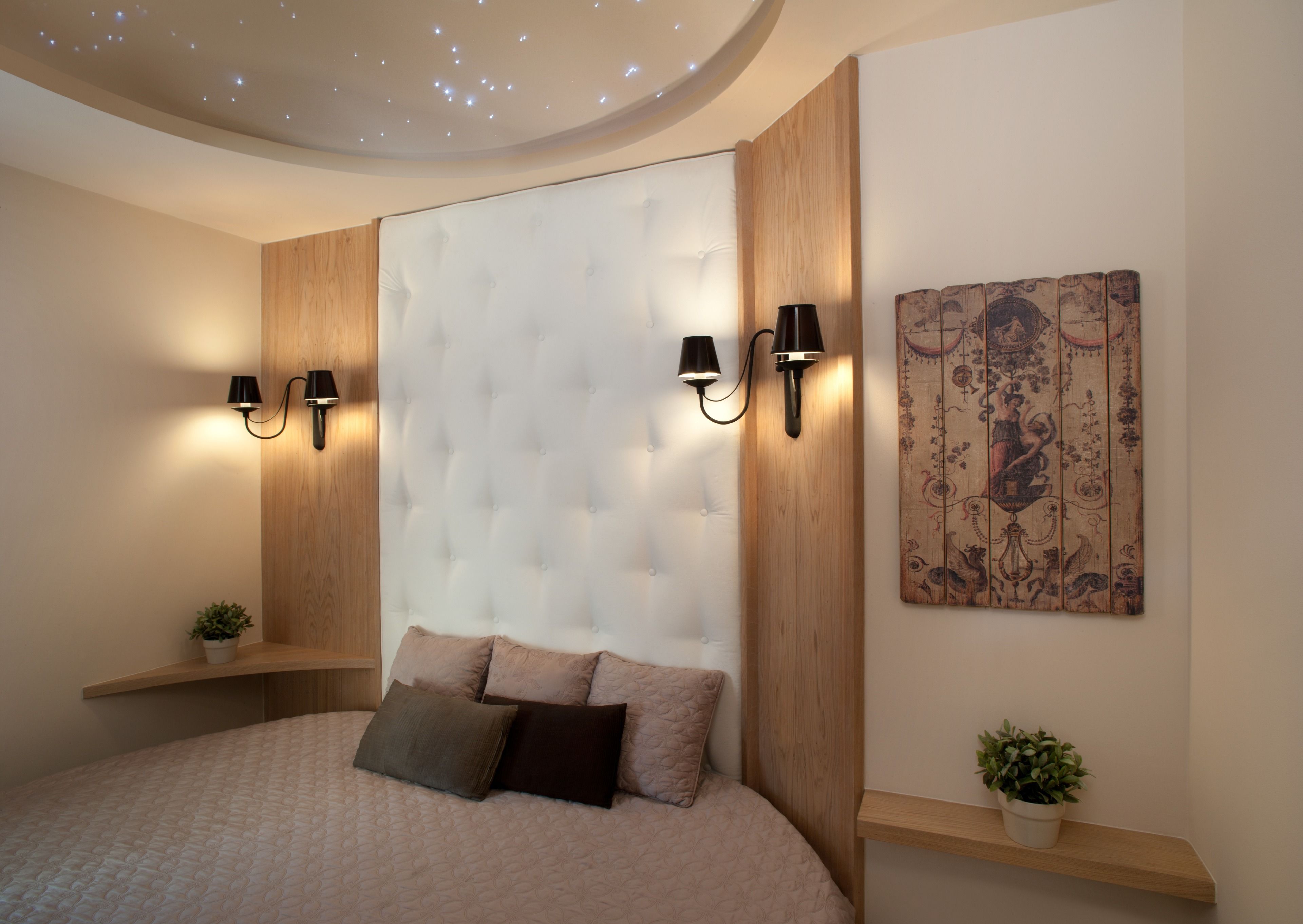 A modern bedroom with a tufted headboard, two wall-mounted lamps, a decorative tapestry, and a bedside plant.