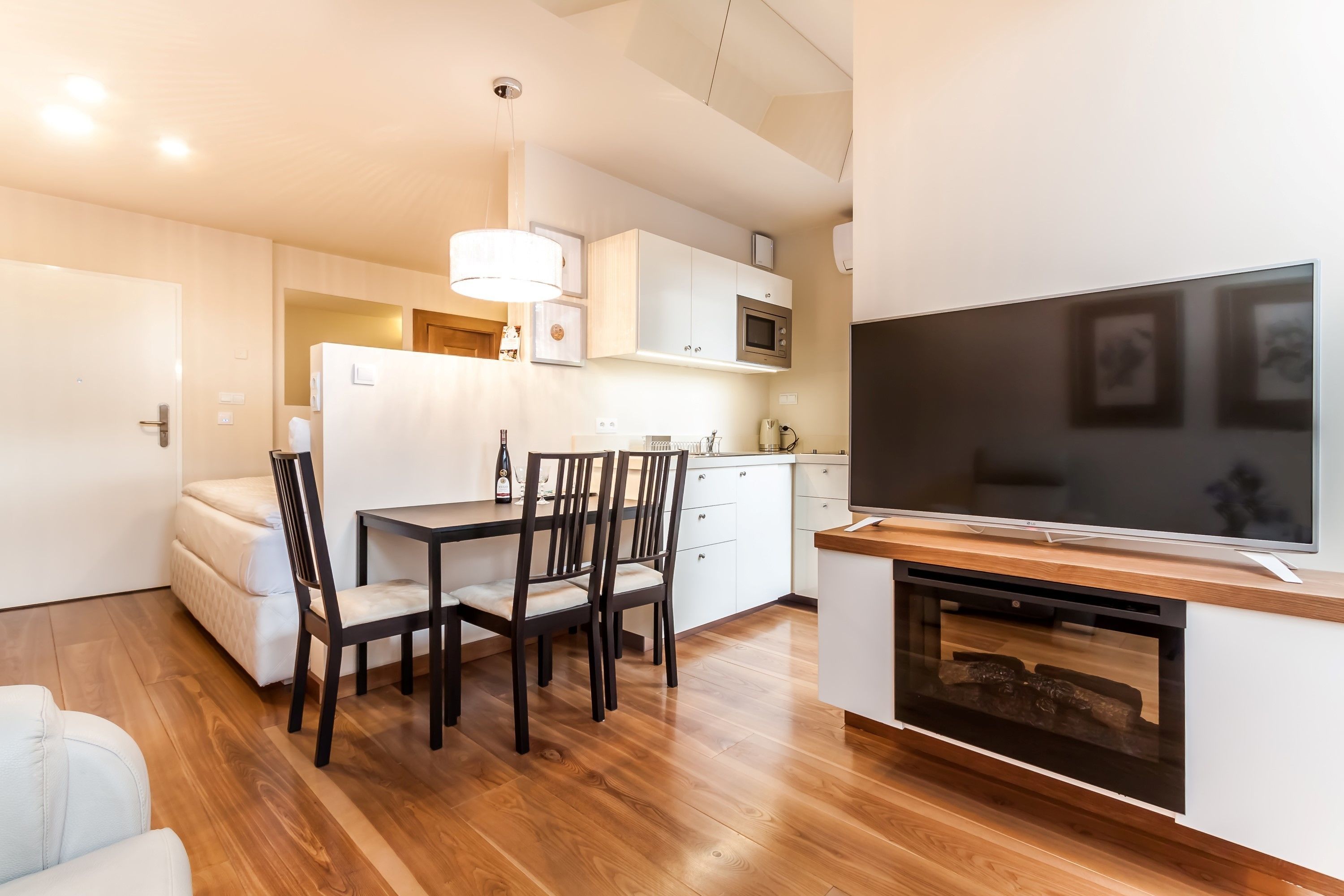Una cocina moderna con armarios blancos, un microondas y un horno empotrado. Cuenta con un comedor con mesa y sillas, y un televisor montado en la pared.