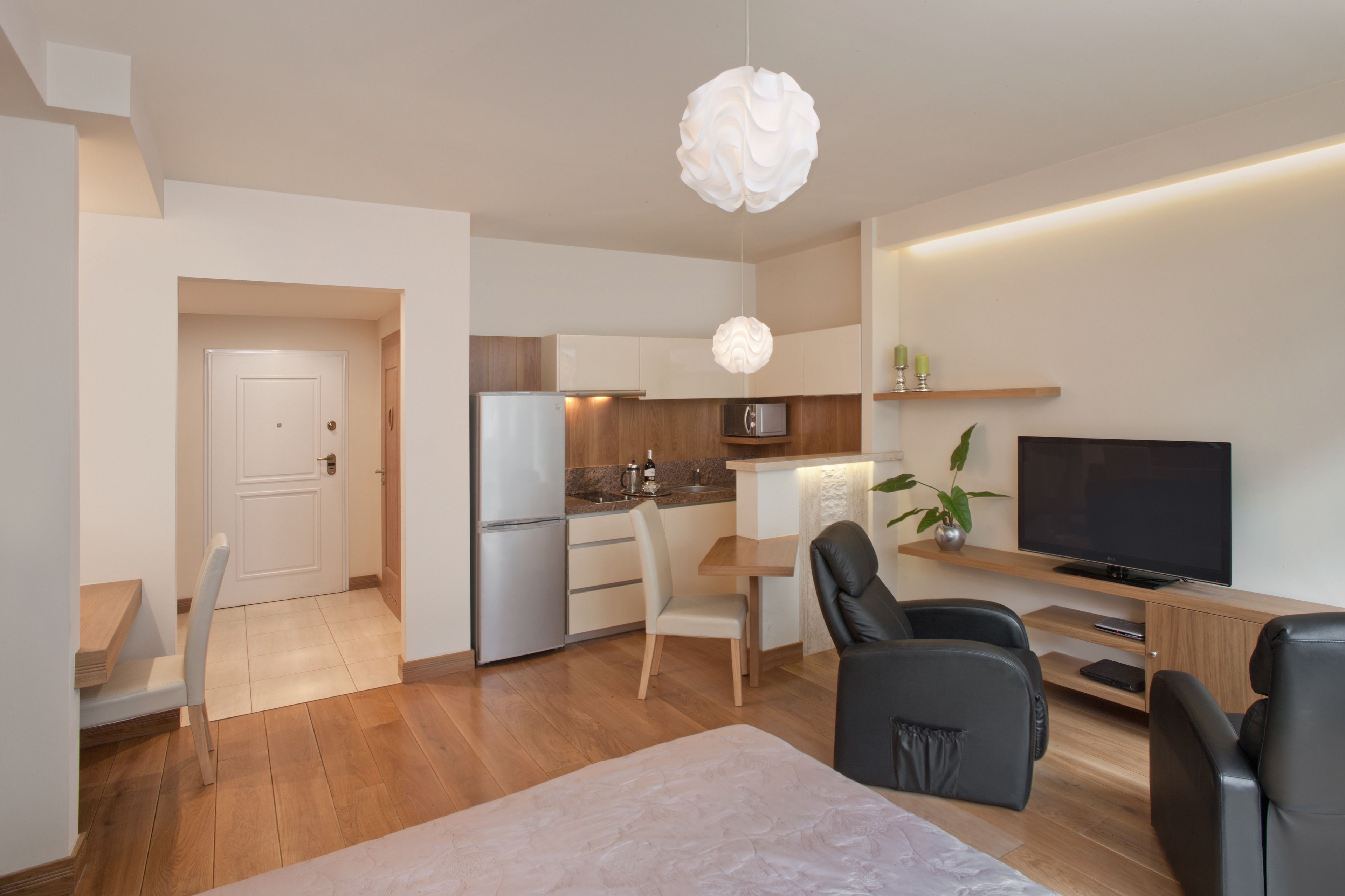 A modern living room with a flat-screen TV, a dining area with a table and chairs, a kitchen with a refrigerator and microwave, and a large white pendant light.
