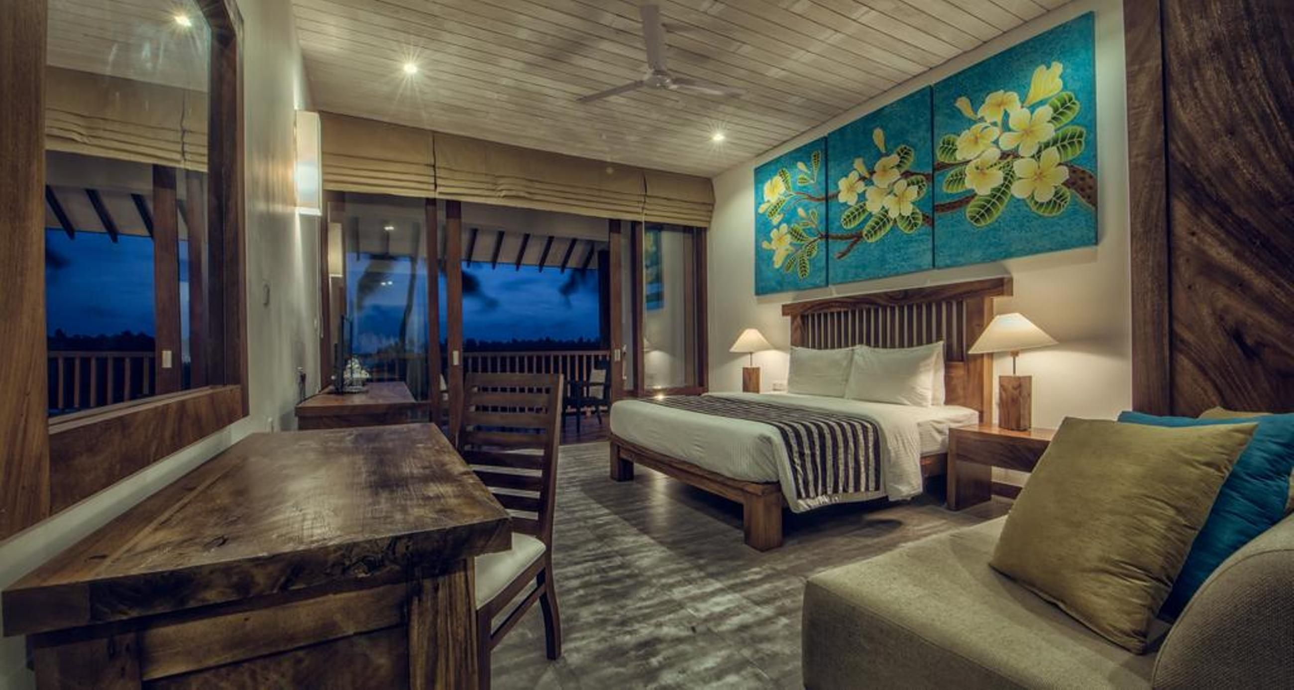 Un dormitorio con cama, mesitas de noche, un escritorio de madera, una silla y un ventanal grande.