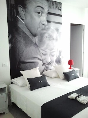 Una camera da letto con un grande poster appeso al muro, un letto con cuscini bianchi e neri, una lampada da comodino rossa e un letto rifatto con copripiumino nero.