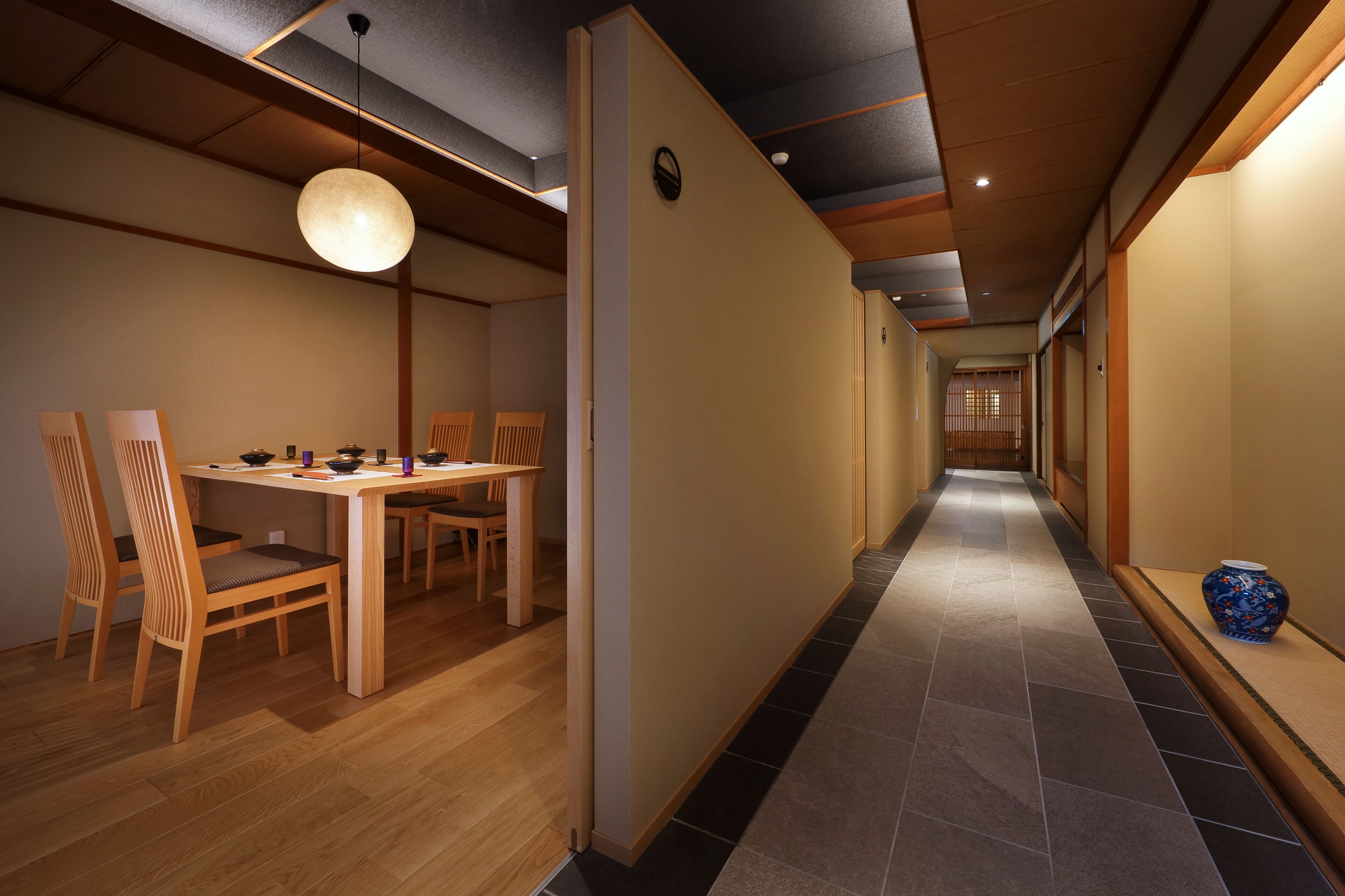 A hallway with a dining area featuring a wooden table and chairs, a hanging light fixture, and a tiled floor.