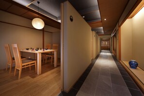 Un couloir qui donne sur un espace repas comprenant une table et des chaises en bois, un luminaire suspendu et un sol carrelé.