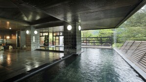 Una piscina interior con suelo de baldosas, zona de terraza de madera y pared de cristal que ofrece vistas a la vegetación exterior.