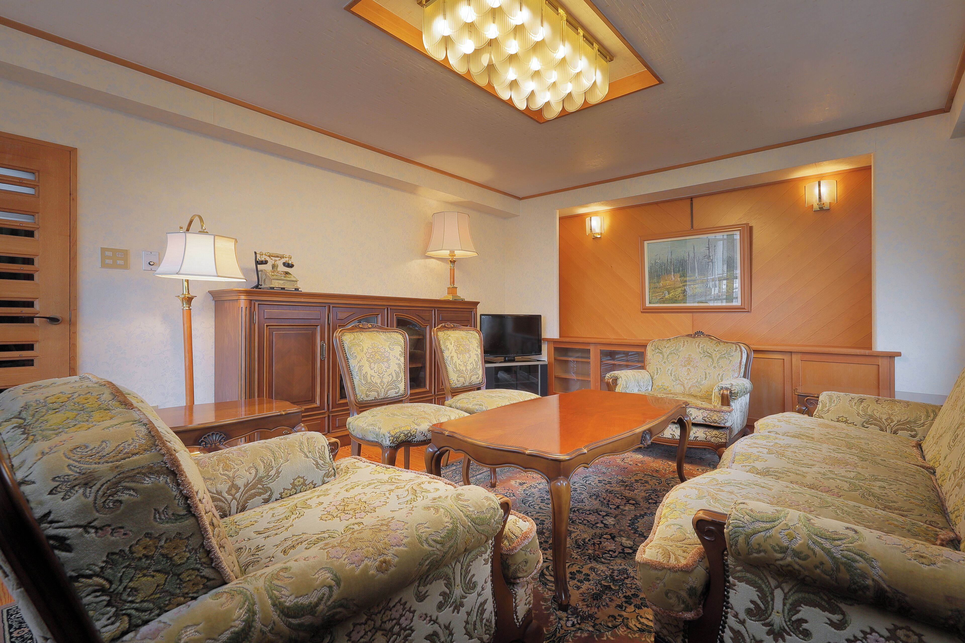 A room with a wooden table, four patterned armchairs, a television, and a painting on the wall.