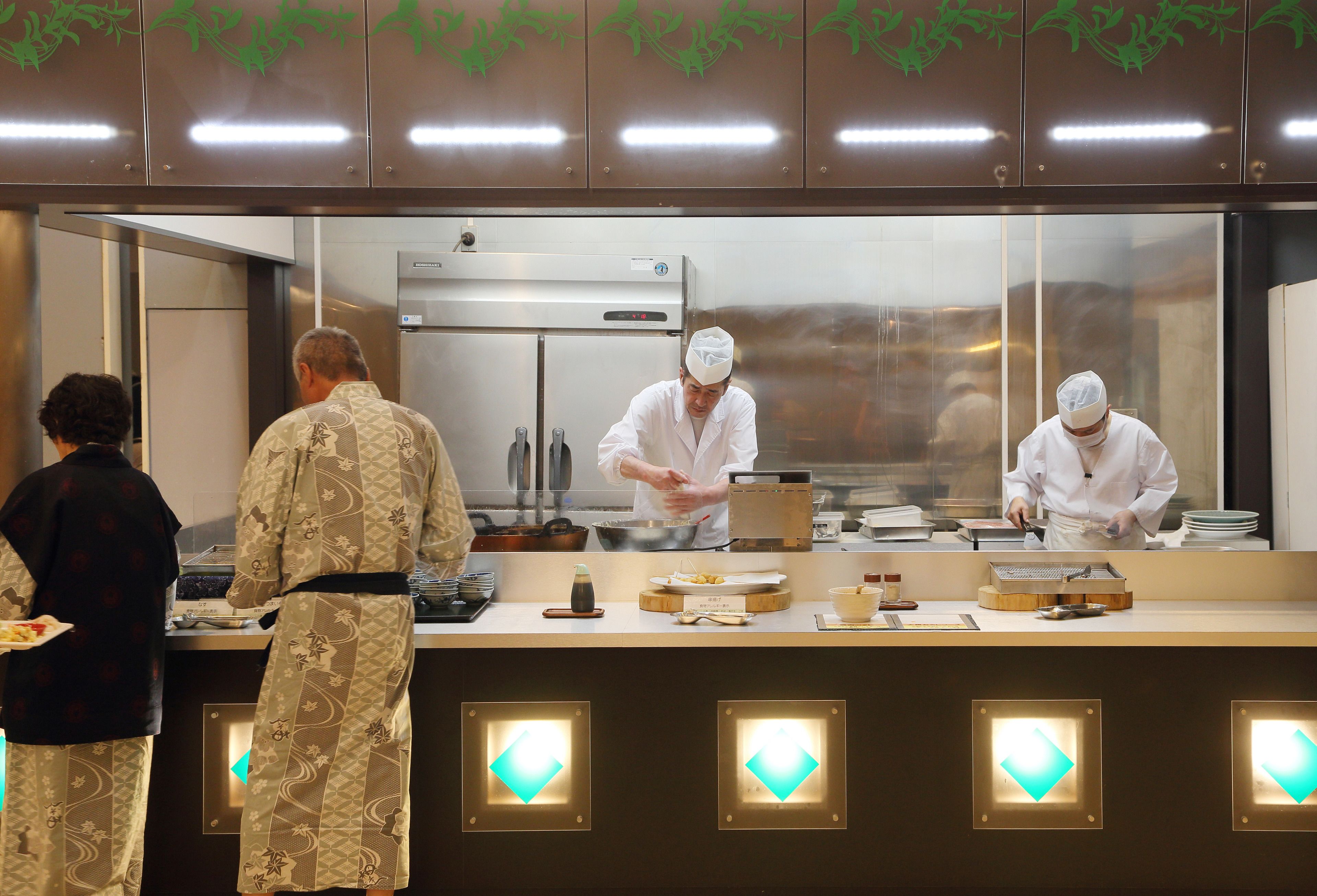 Un espace cuisine équipé d’appareils électroménagers en acier inoxydable, un comptoir avec des aliments et deux personnes portant des vêtements traditionnels.