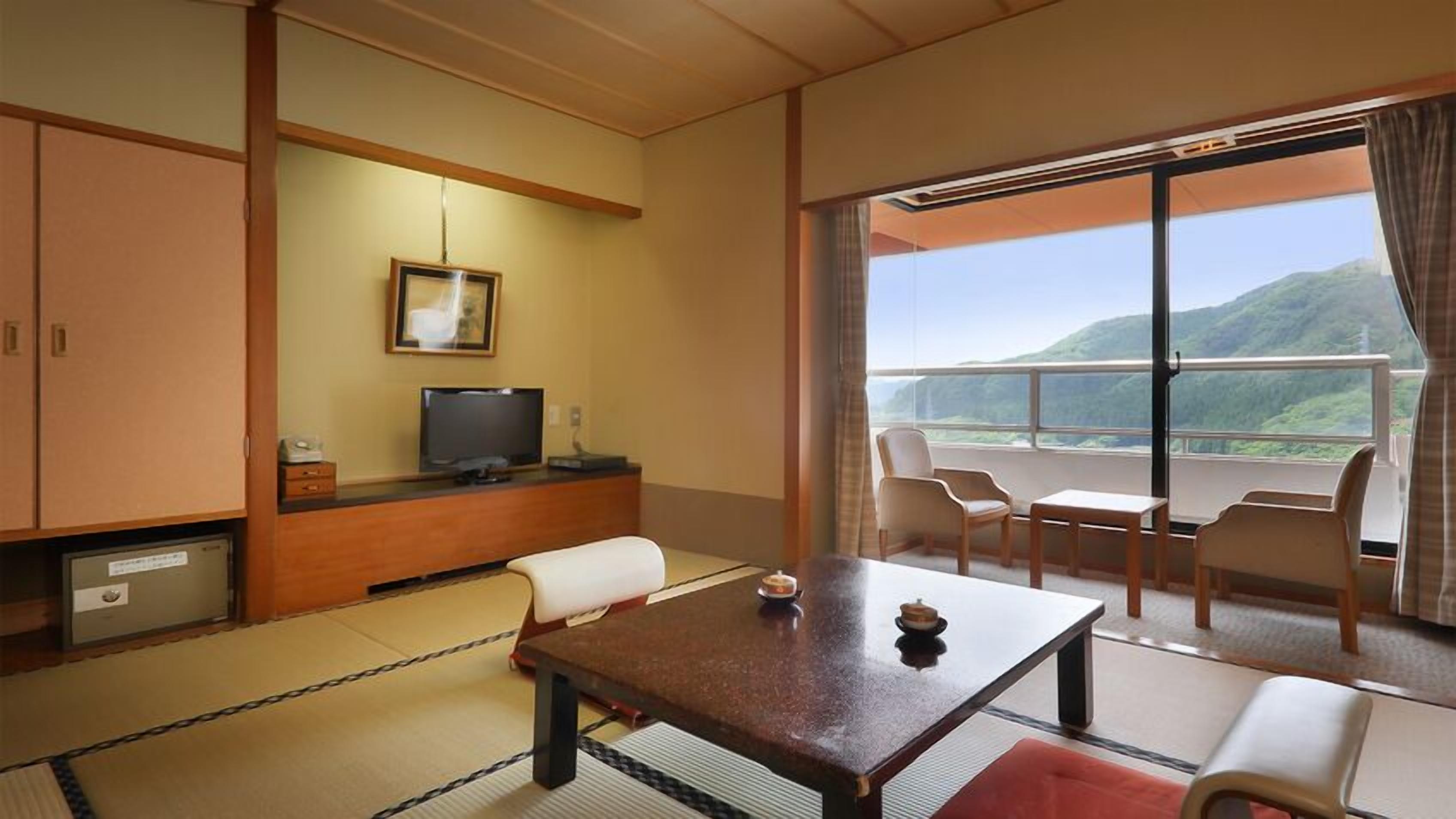 A hotel room with tatami flooring, a television, a dining table, and a balcony with a view of hills.