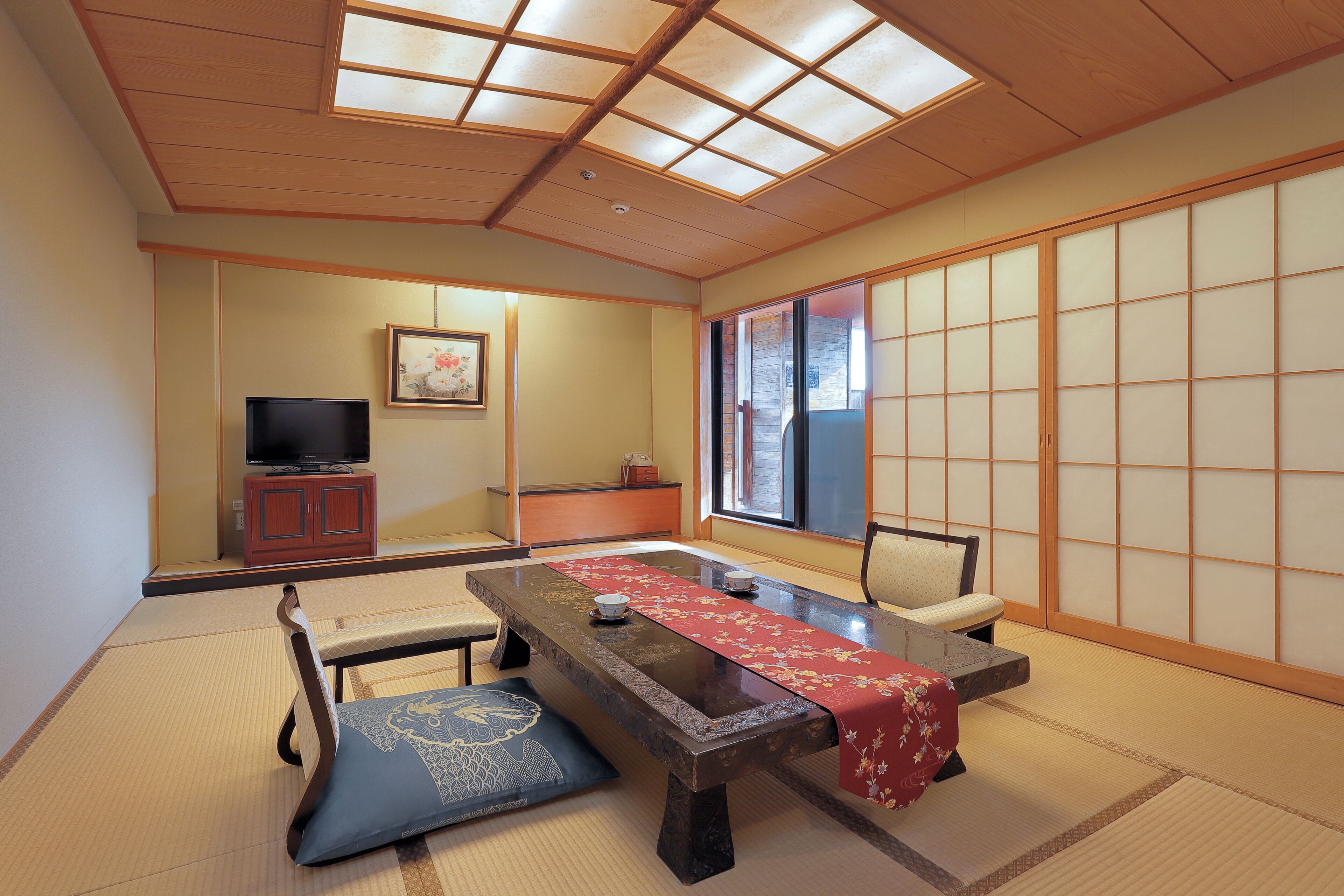 Una habitación japonesa tradicional con suelo de tatami, una mesa baja con mantel rojo, un televisor sobre un mueble y puertas correderas.
