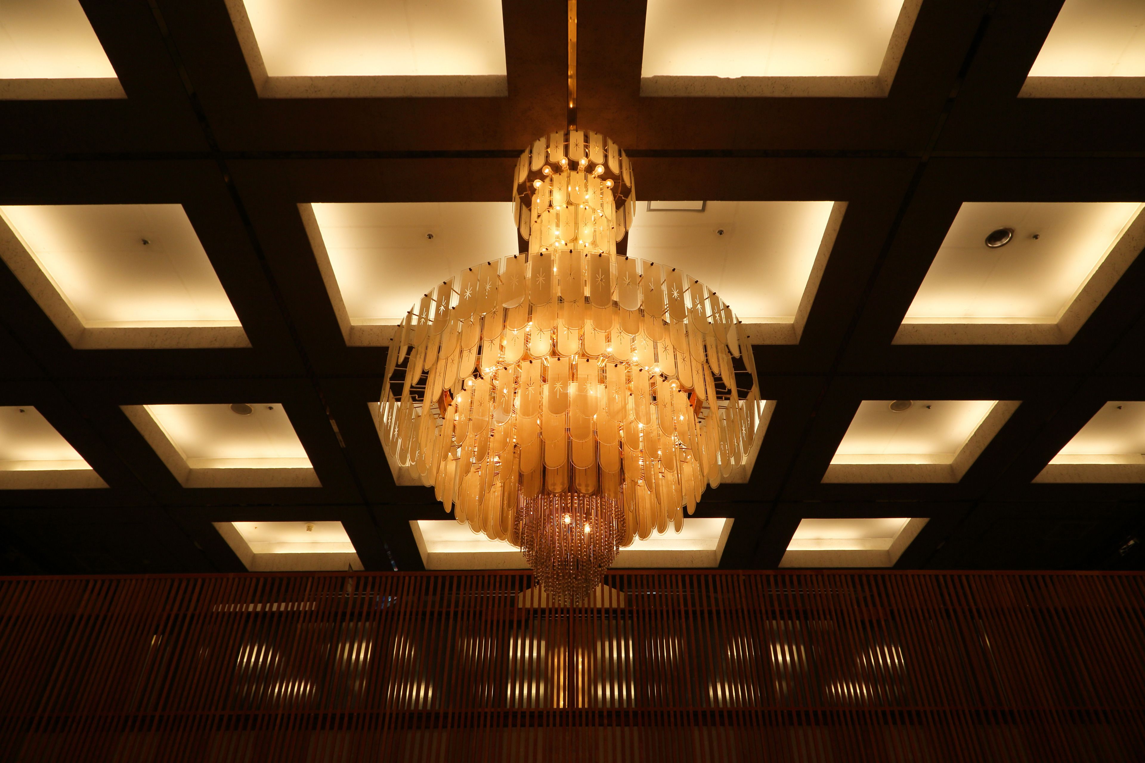 En kristallkrona med flera våningar och ljuskällor, hängande från ett tak med infälld belysning.