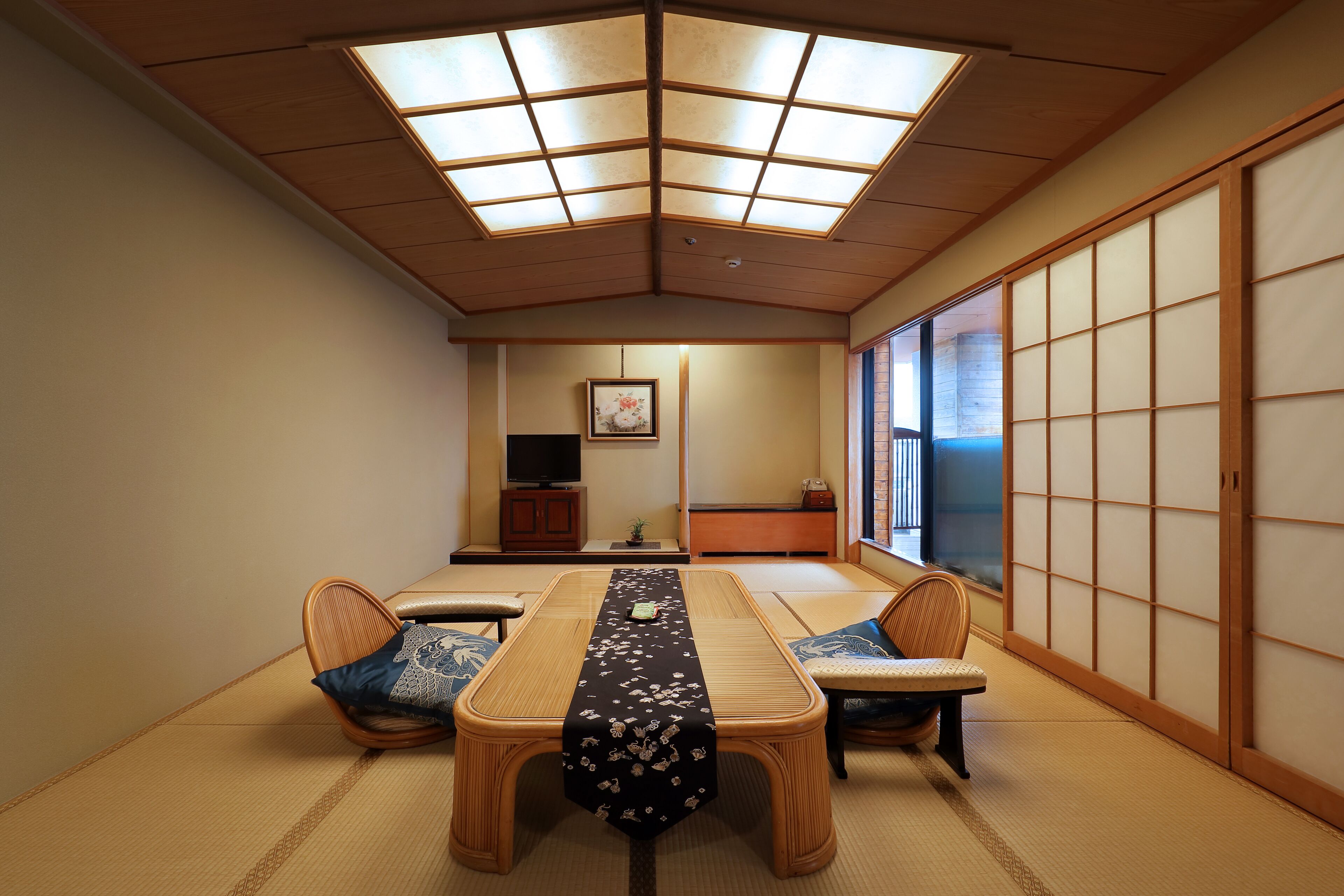 A traditional Japanese room with tatami flooring, a low table with cushions, sliding doors, and a television in the background.
