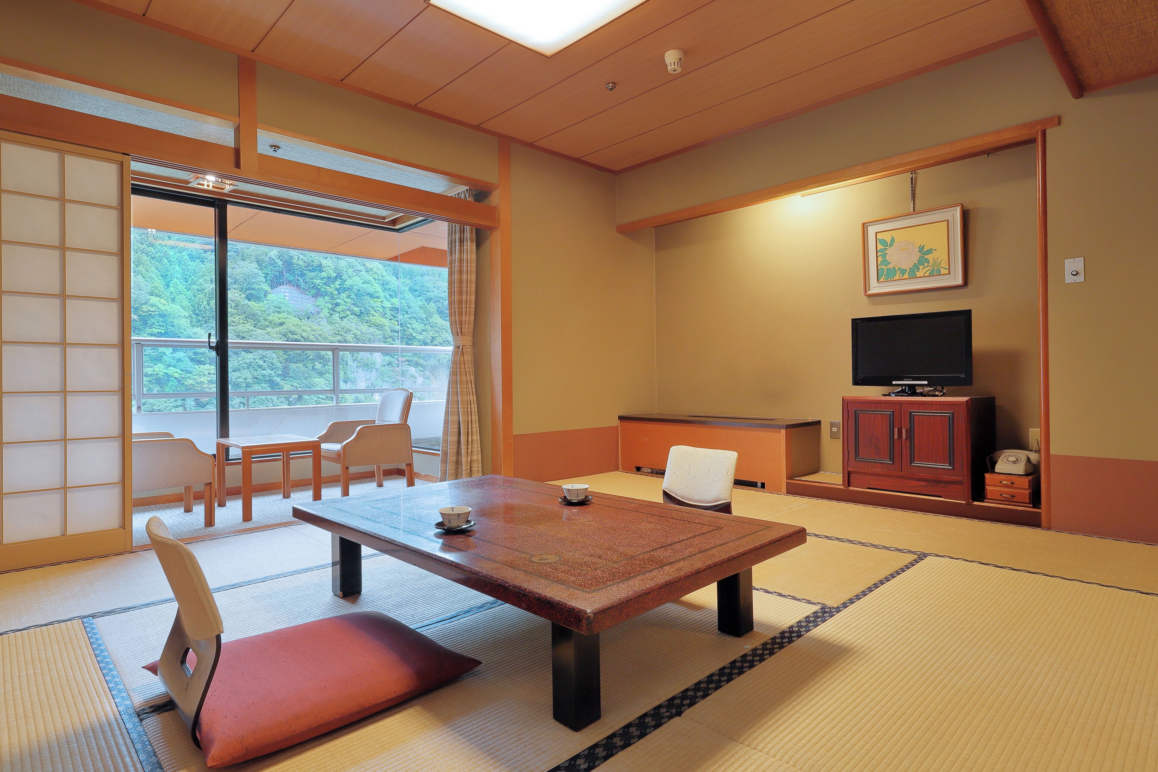 Una habitación japonesa tradicional con suelo de tatami, una mesa baja con cojines, un televisor y puertas correderas que dan a un balcón.