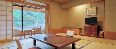 Una habitación japonesa tradicional con suelo de tatami, una mesa baja con cojines, un televisor y puertas correderas que dan a un balcón.