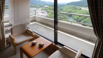 A balcony with two chairs, a small table, and a view of a green landscape with mountains and a river.