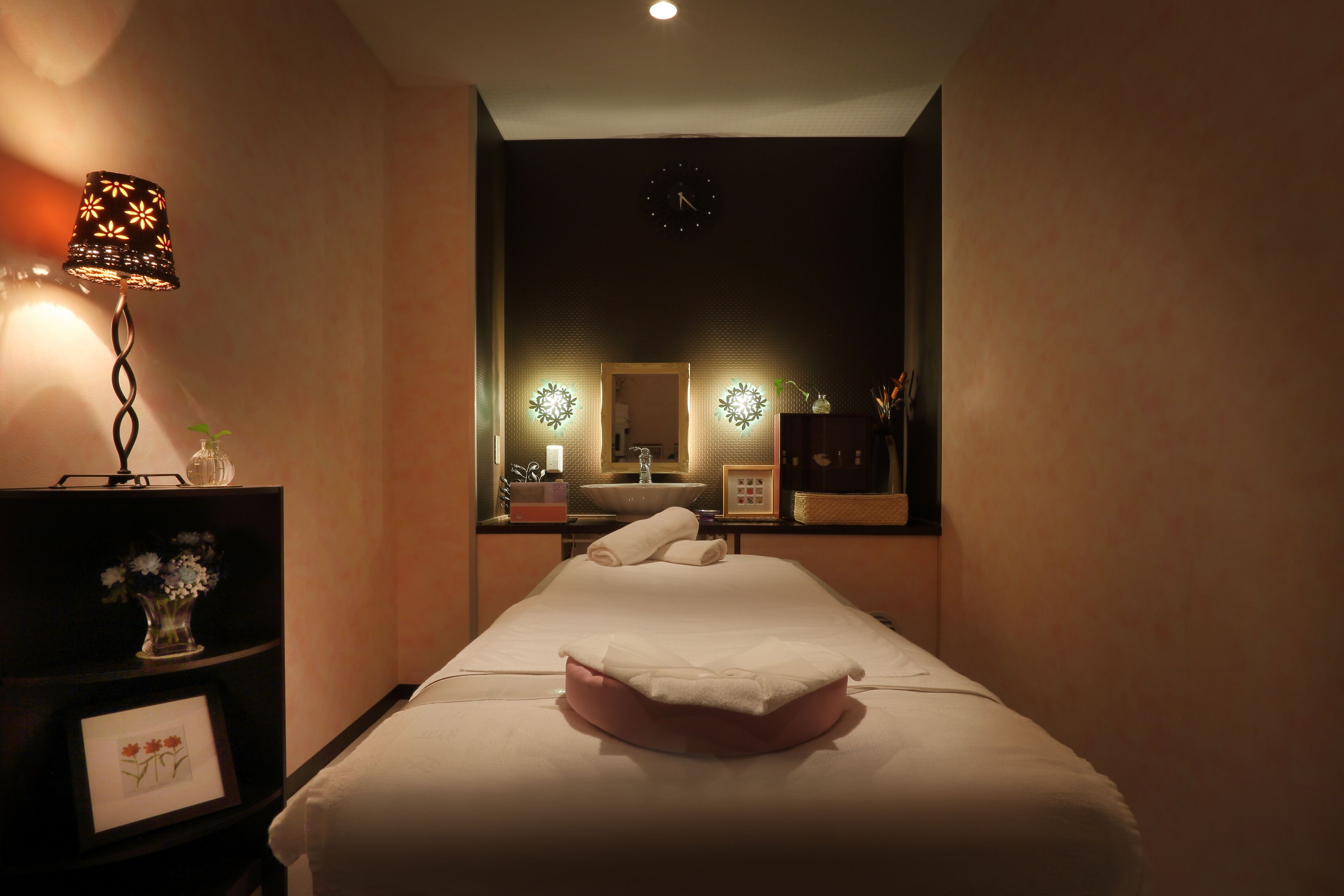 Una habitación tenuemente iluminada con una cama, una lámpara de noche, un espejo y una repisa con un florero y un marco de fotos.