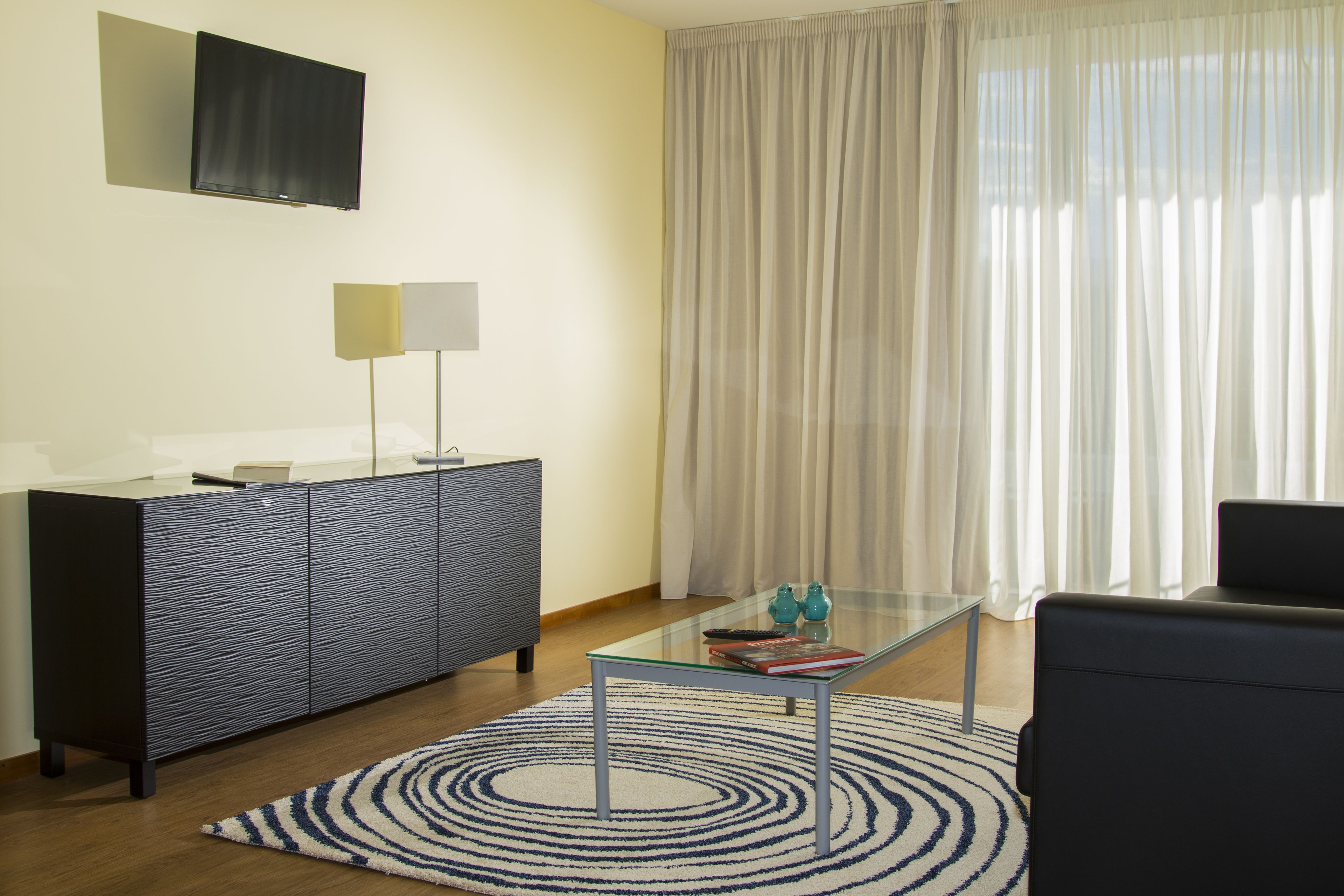 Una sala moderna con una mesa de centro de vidrio, un sofá de color oscuro, un tapete estampado, un mueble con un televisor de pantalla plana montado encima y cortinas traslúcidas en la ventana.