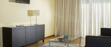 Una sala moderna con una mesa de centro de vidrio, un sofá de color oscuro, un tapete estampado, un mueble con un televisor de pantalla plana montado encima y cortinas traslúcidas en la ventana.