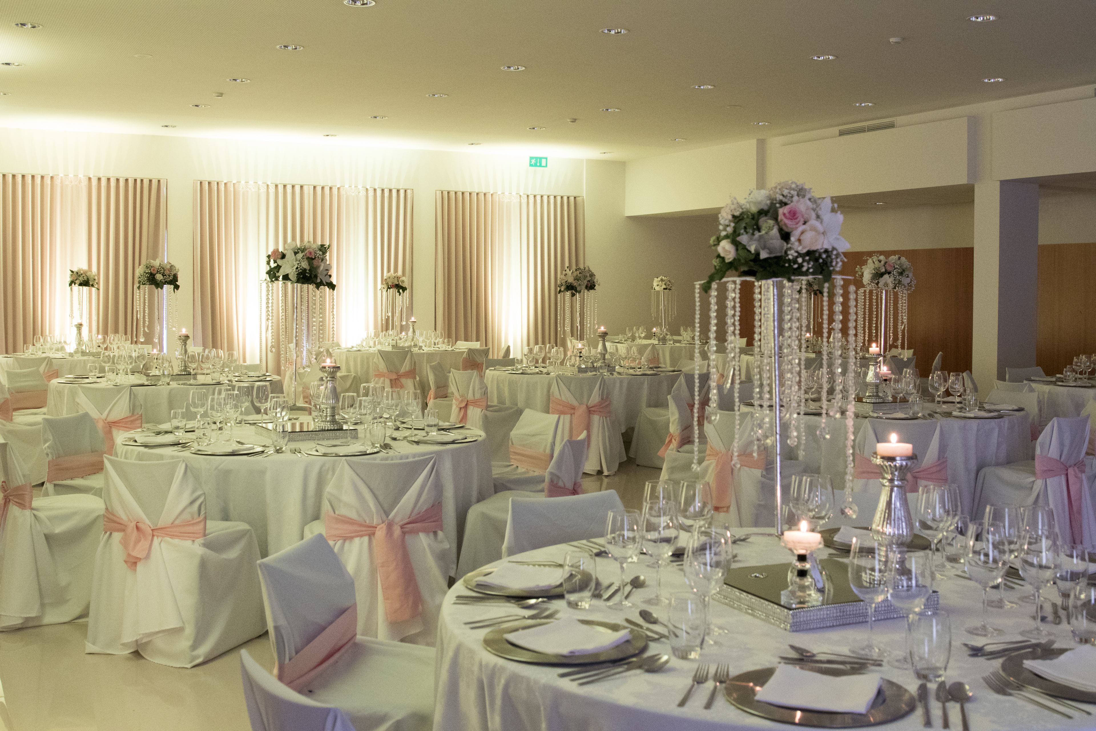 Une salle de banquet avec des tables rondes dressées pour le repas, chacune étant recouverte d’une nappe blanche et ornée d’un ruban rose.