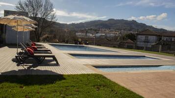 Una piscina con sillas de descanso y sombrillas, rodeada de césped y edificios al fondo.