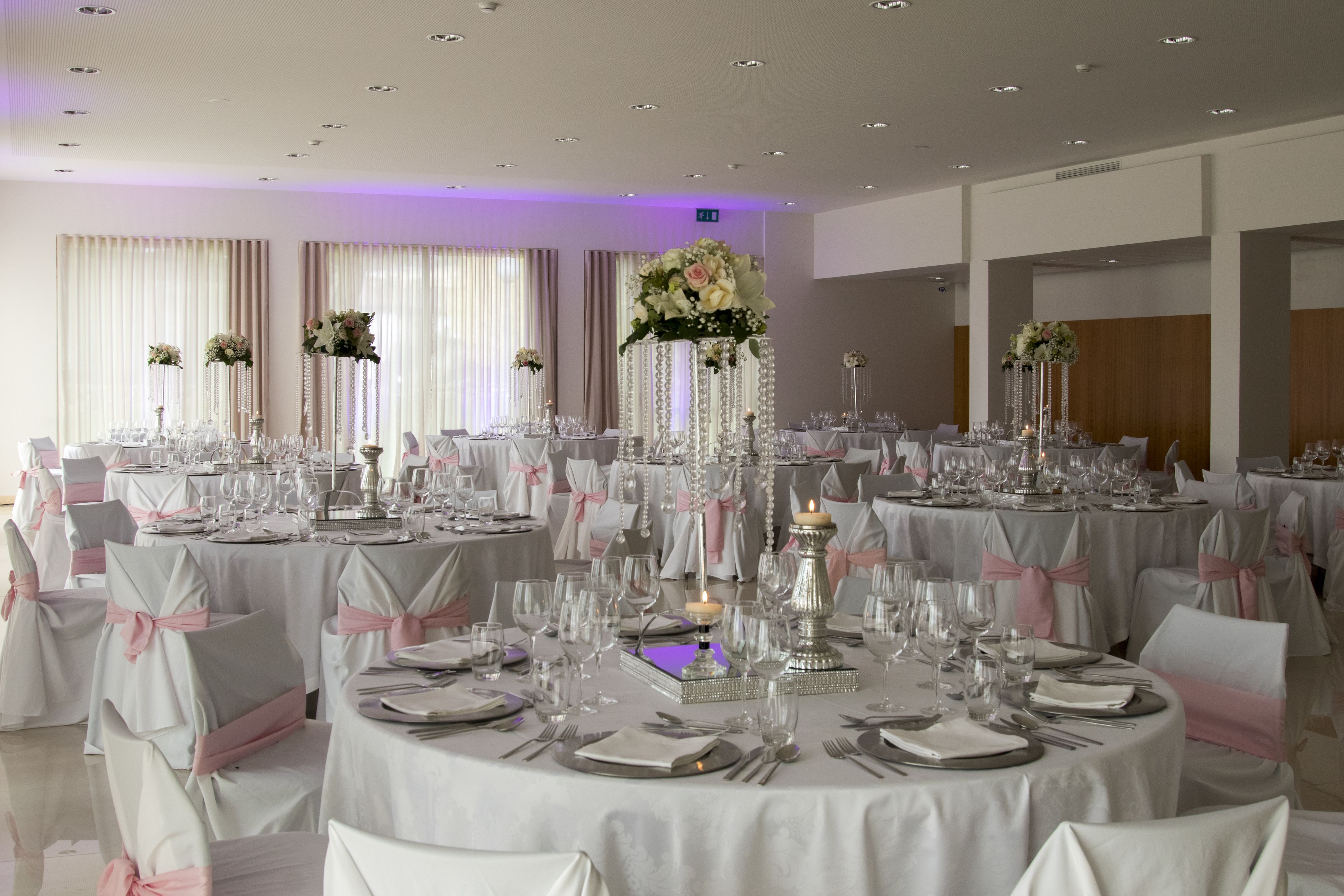 Un salón de banquetes con mesas redondas dispuestas para un evento formal, cada una decorada con manteles blancos, cintas rosas y centros de mesa florales.