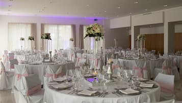 Un salón de banquetes con mesas redondas dispuestas para un evento formal, cada una decorada con manteles blancos, cintas rosas y centros de mesa florales.