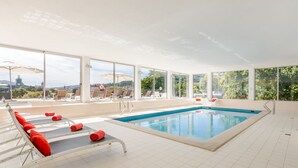 An indoor swimming pool with sun loungers and umbrellas, large windows offering a view of the outdoors.