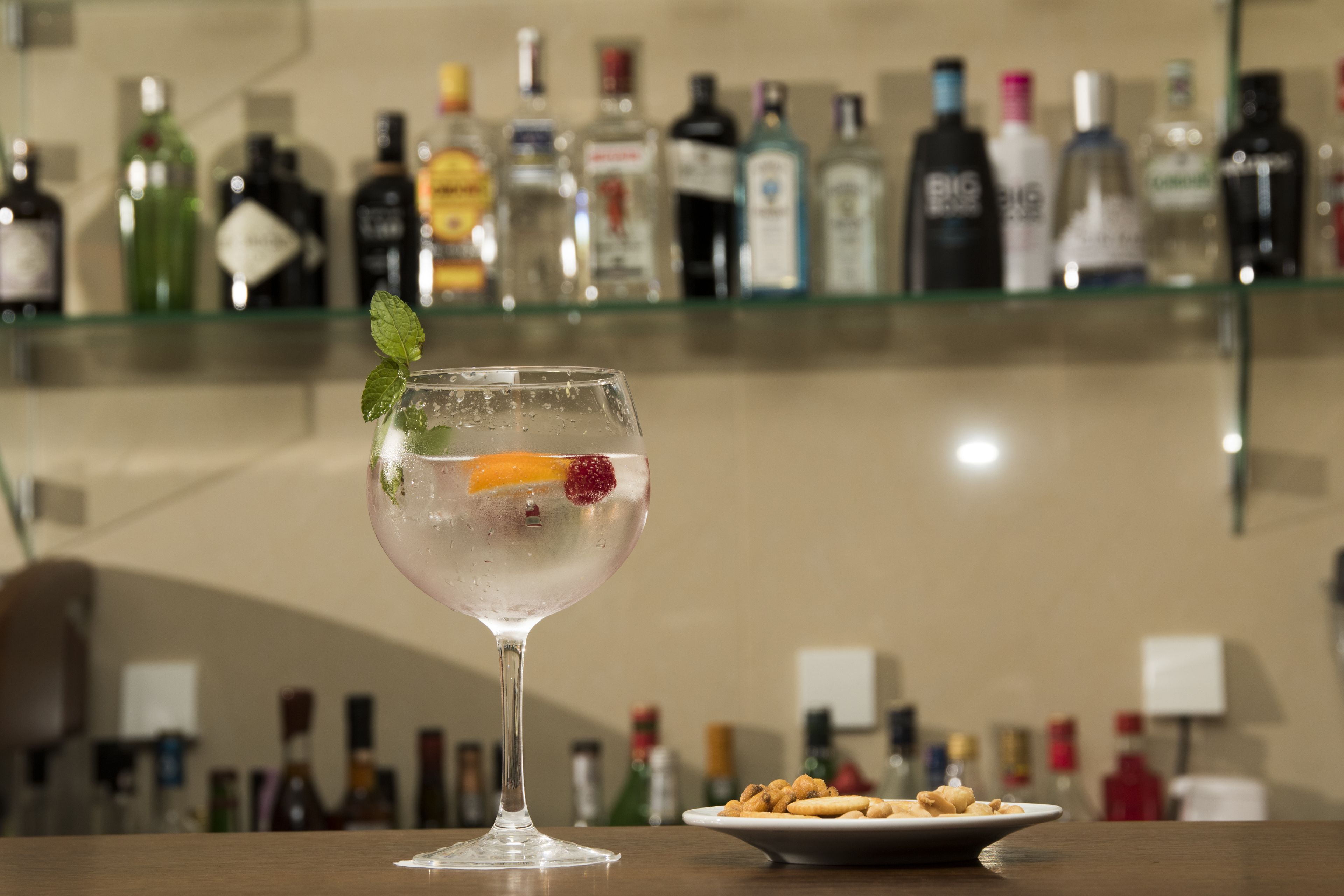 A bar area with a glass of a clear cocktail garnished with a mint leaf and an orange slice, a plate of snacks, and a shelf with various liquor bottles in the background.