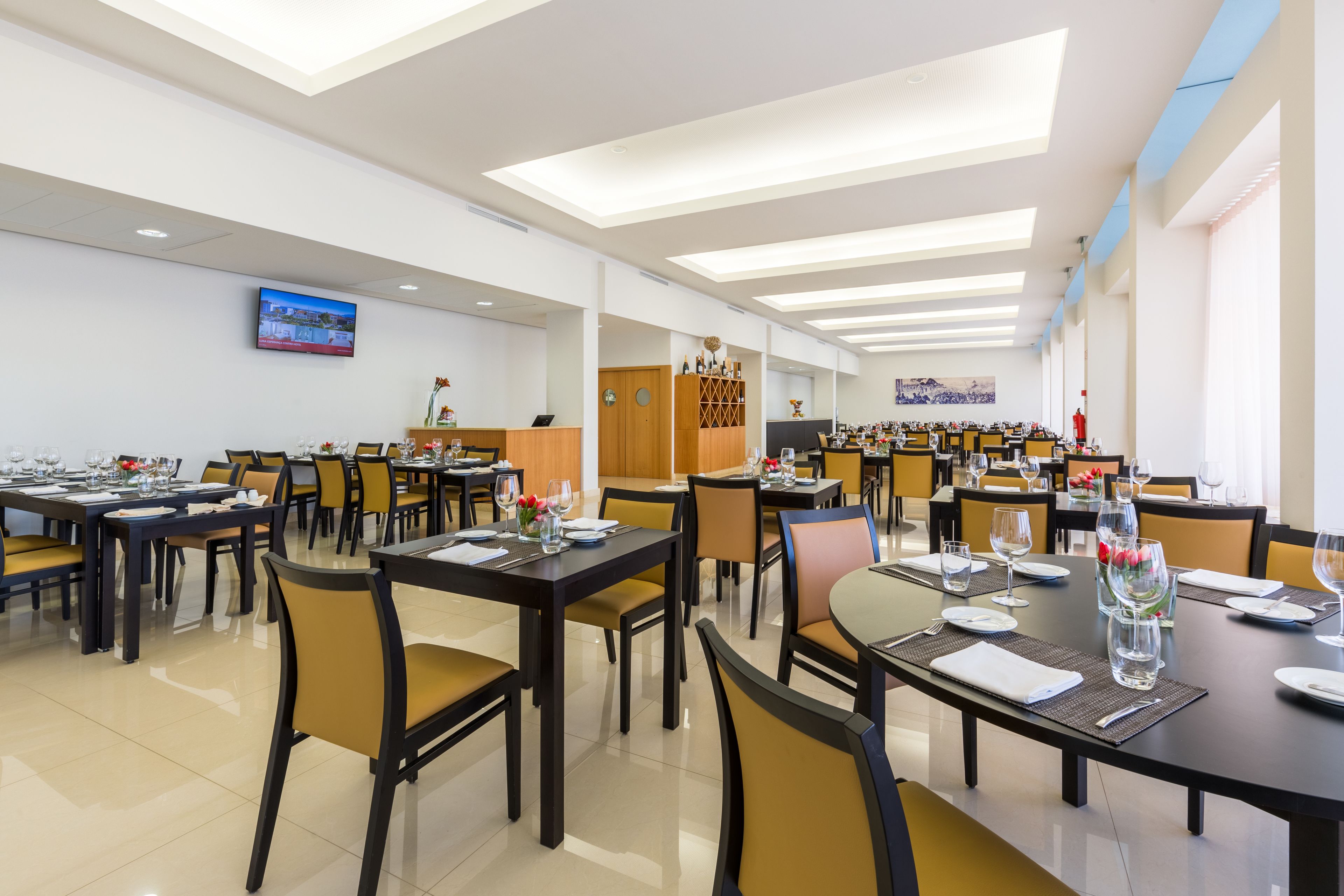 Un vaste espace de restauration dans l’hôtel, avec de nombreuses tables et chaises, un téléviseur à écran plat fixé au mur et une grande fenêtre laissant entrer la lumière naturelle.