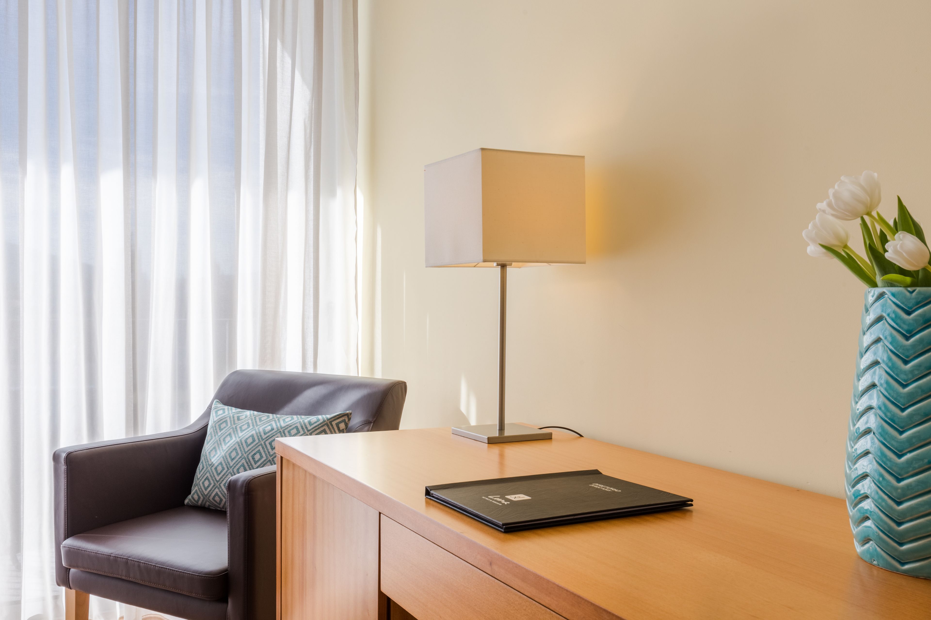 A hotel room with a wooden desk, a modern lamp, a laptop, a chair with a patterned pillow, and a vase with white flowers.