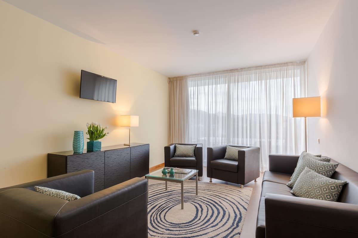 Una sala de estar moderna con un sofá, dos sillones, una mesa de centro, un televisor de pantalla plana montado en la pared y un amplio ventanal con cortinas traslúcidas.