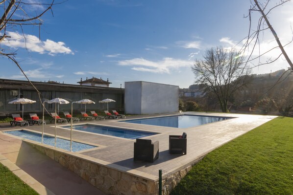 Piscina rectangular con sillas de descanso y sombrillas, rodeada por una terraza de piedra y una zona verde.