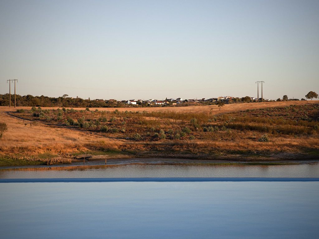 A rural landscape with a body of water, dry grassland, and a few scattered buildings in the distance.