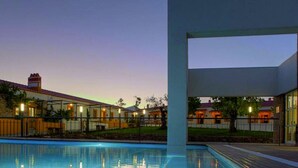 A pool area with a modern building in the background, a clear sky, and a well-lit patio. - Herdade da Urgueira (Vila Velha de Rodao)