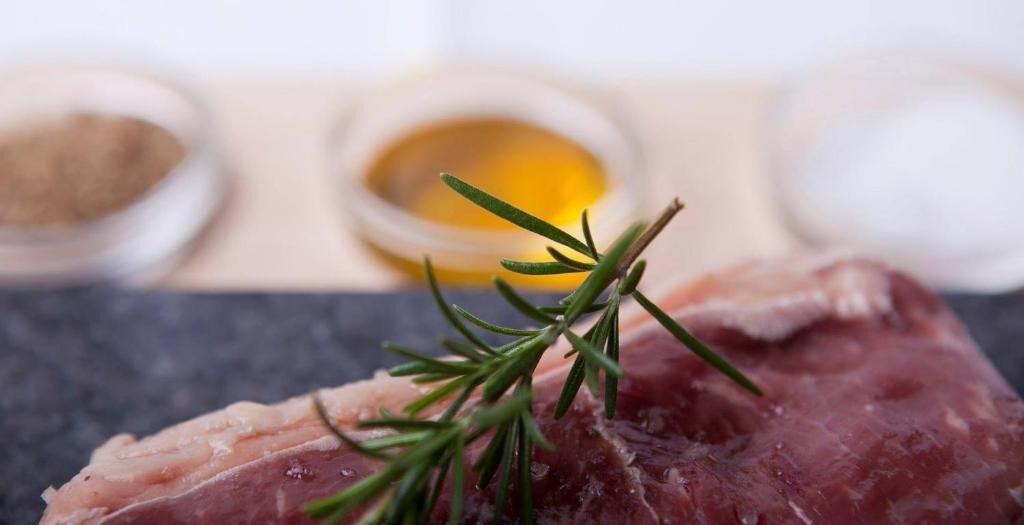 Primer plano de carne cruda con romero y dos recipientes pequeños de condimentos y aceite.