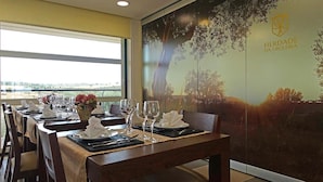 A dining area with tables set for a meal, a large window, and a wall with a tree and 'Herdade da Urgêira' logo. - Herdade da Urgueira (Vila Velha de Rodao)