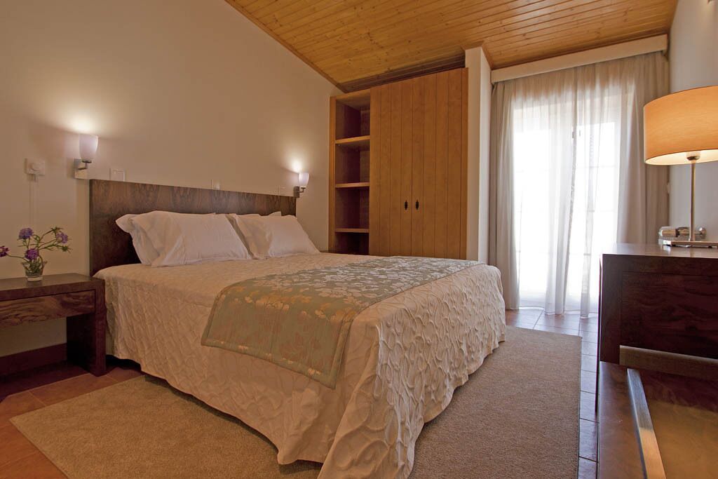 Un dormitorio con cabecera de madera, cama con sábanas blancas, armario de madera, mesita de noche con lámpara y ventana con cortinas.