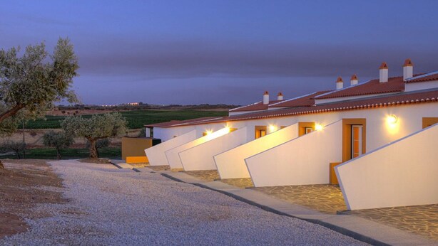 A row of modern, white buildings with red-tiled roofs, arranged in a linear fashion along a gravel path. - Herdade da Urgueira (Vila Velha de Rodao)