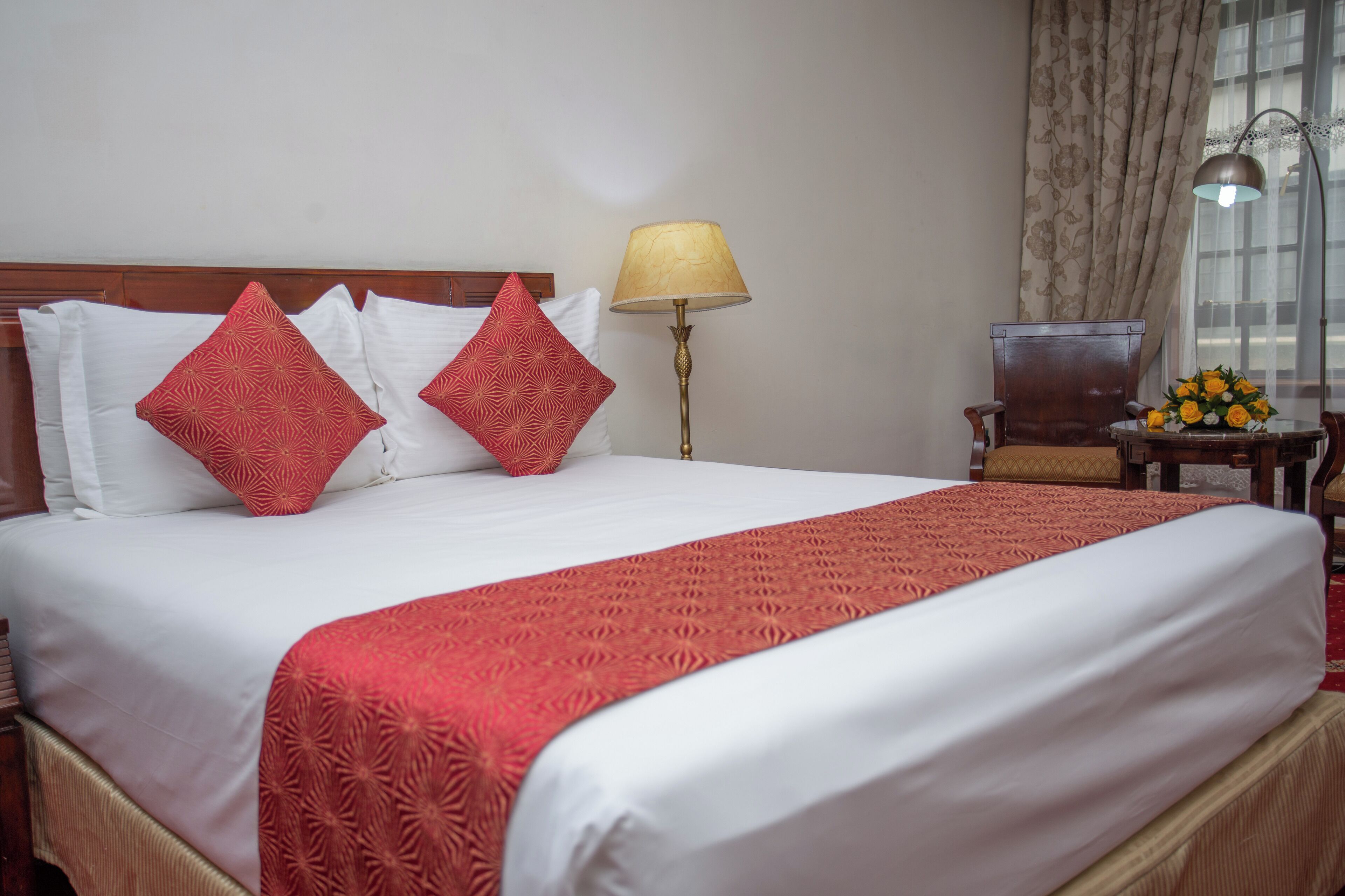 A hotel room with a large bed, a wooden headboard, a lamp, a chair, and a table with a vase of flowers.