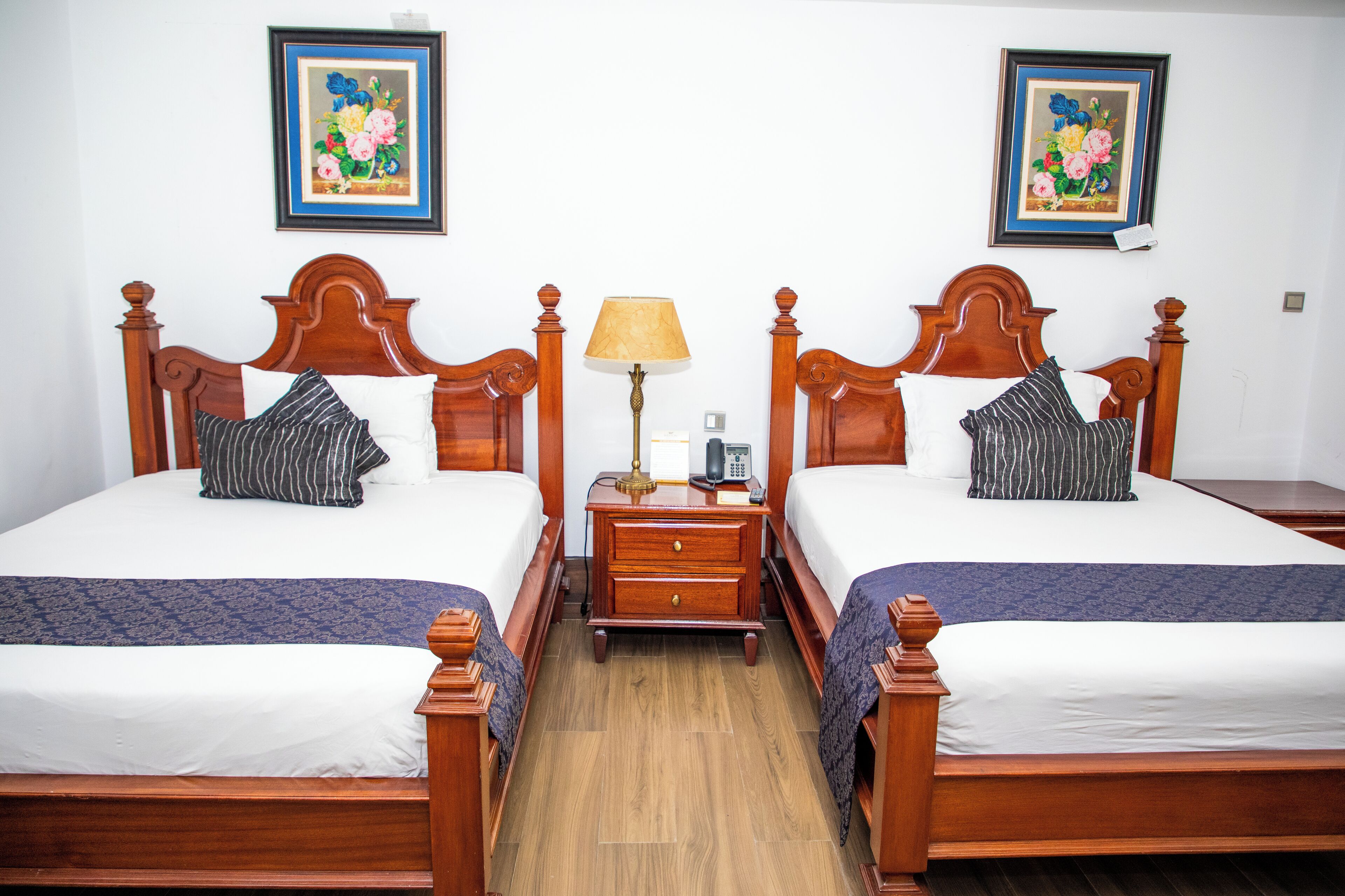 Habitación de hotel con dos camas de madera, una mesita de noche con lámpara y dos cuadros enmarcados con motivos florales en la pared.