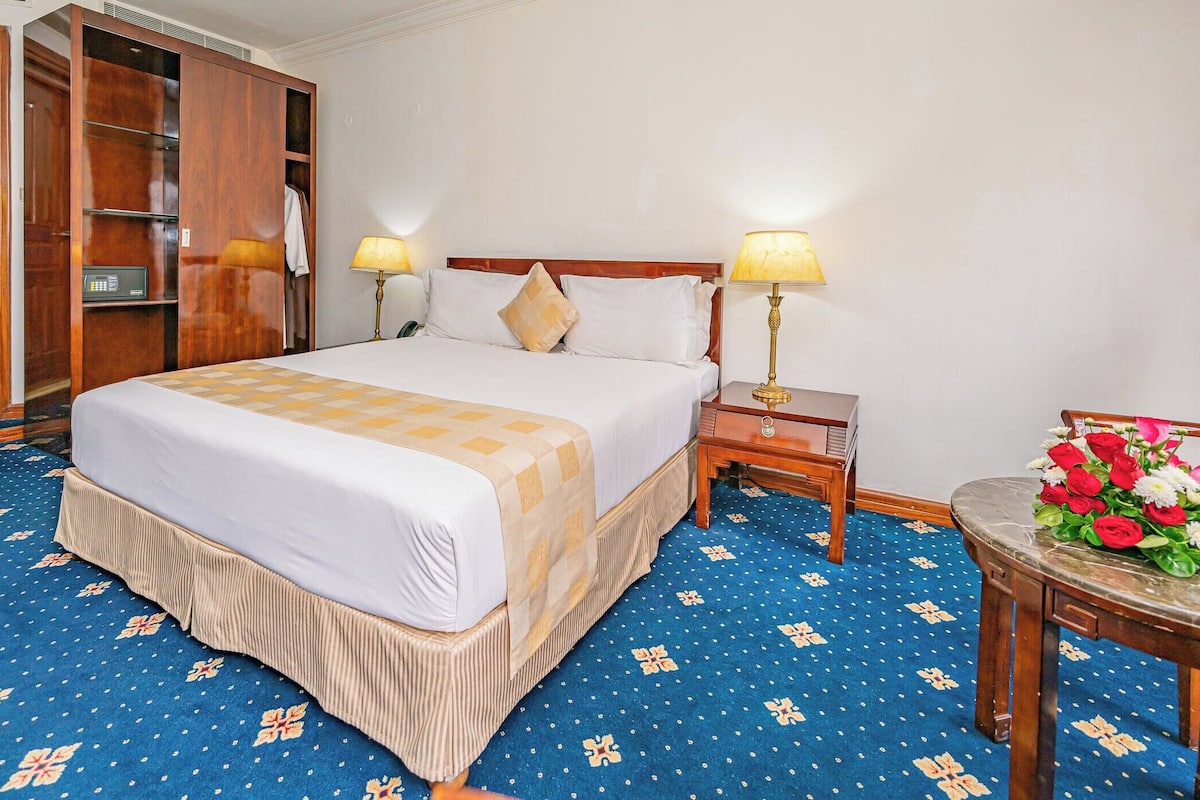 Habitación de hotel con una cama grande, dos lámparas de noche, un armario de madera y una pequeña mesa con un ramo de flores.