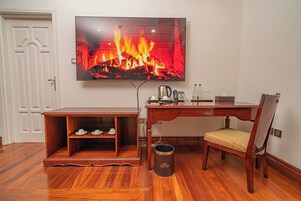 A room with a TV displaying a fireplace, a wooden desk, a chair, and a small cabinet with shoes.