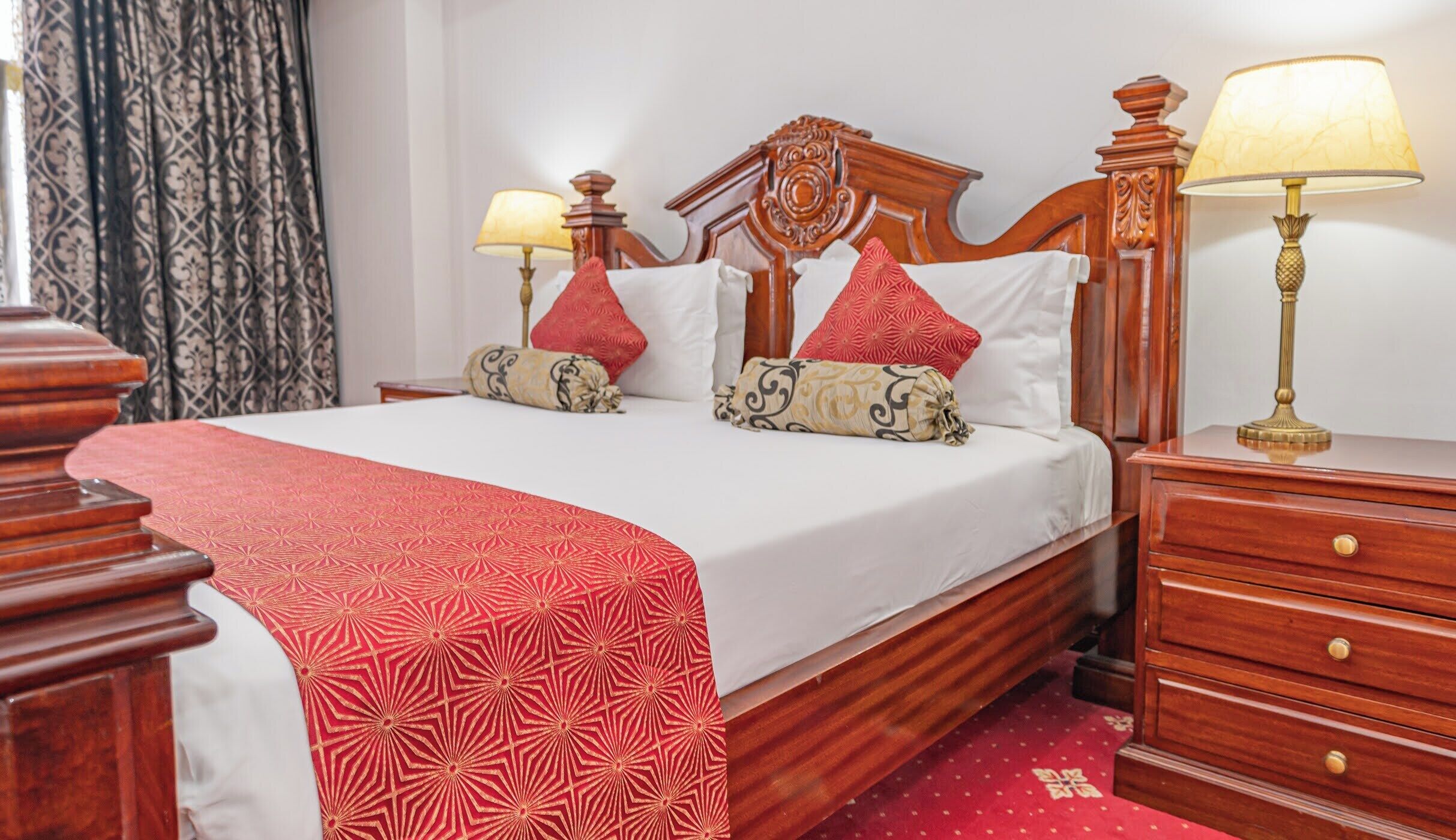 A neatly made bed with a red patterned bedspread, two bedside lamps, and a wooden nightstand with drawers.