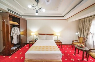 Una habitación de hotel con una cama grande, dos mesitas de noche con lámparas, un armario de madera, piso cubierto de alfombra roja y un candelabro.