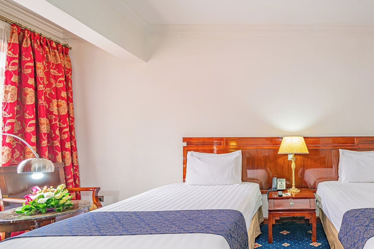 Habitación de hotel con dos camas, cabecero de madera, mesita de noche con lámpara, teléfono y armario.
