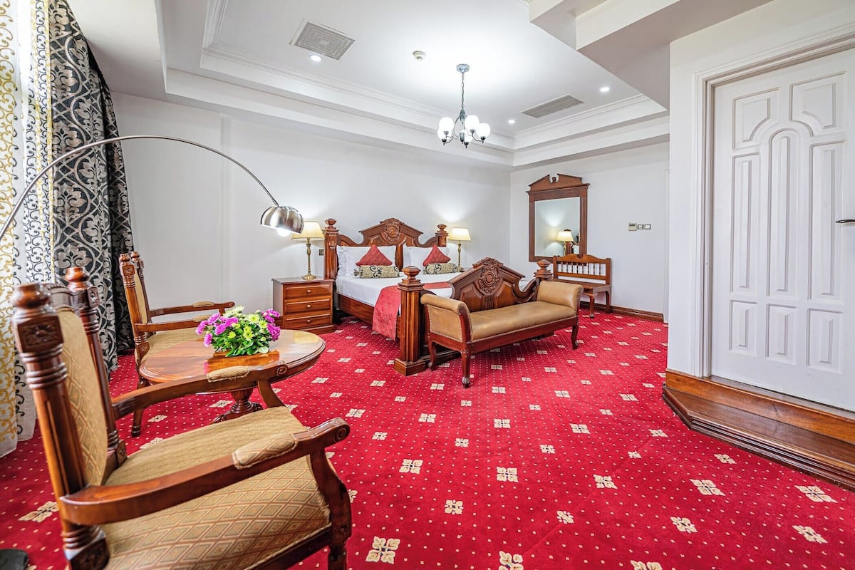 Una habitación de hotel con alfombra de motivos rojos, un sofá y una silla de madera, una pequeña mesa redonda con un jarrón de flores, una cama con cabecero de madera, una mesita de noche con una lámpara, un espejo y una puerta blanca.