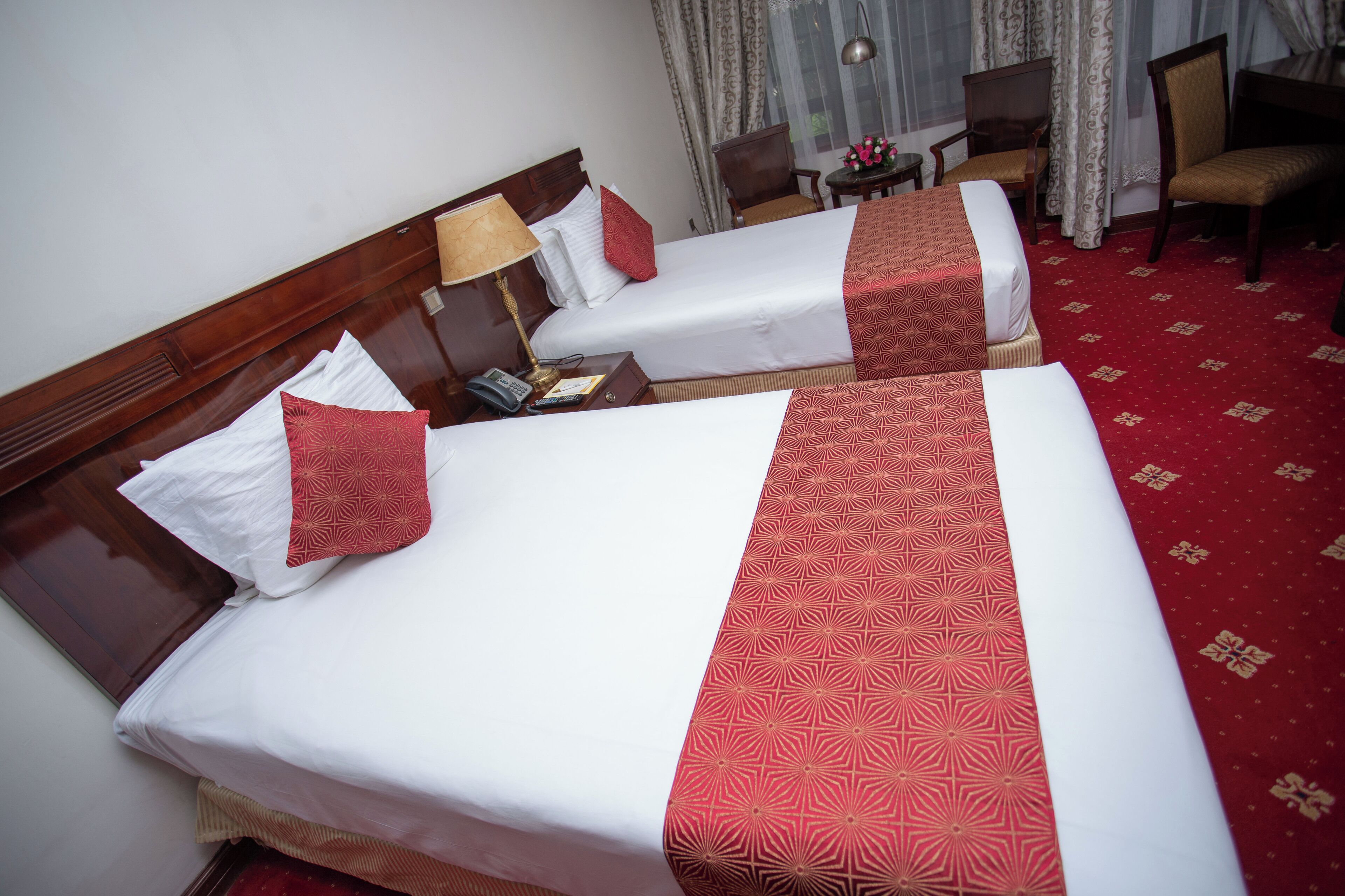 Two neatly made beds with white linens and red decorative pillows, a wooden headboard, a small table with a lamp, and a red carpeted floor.