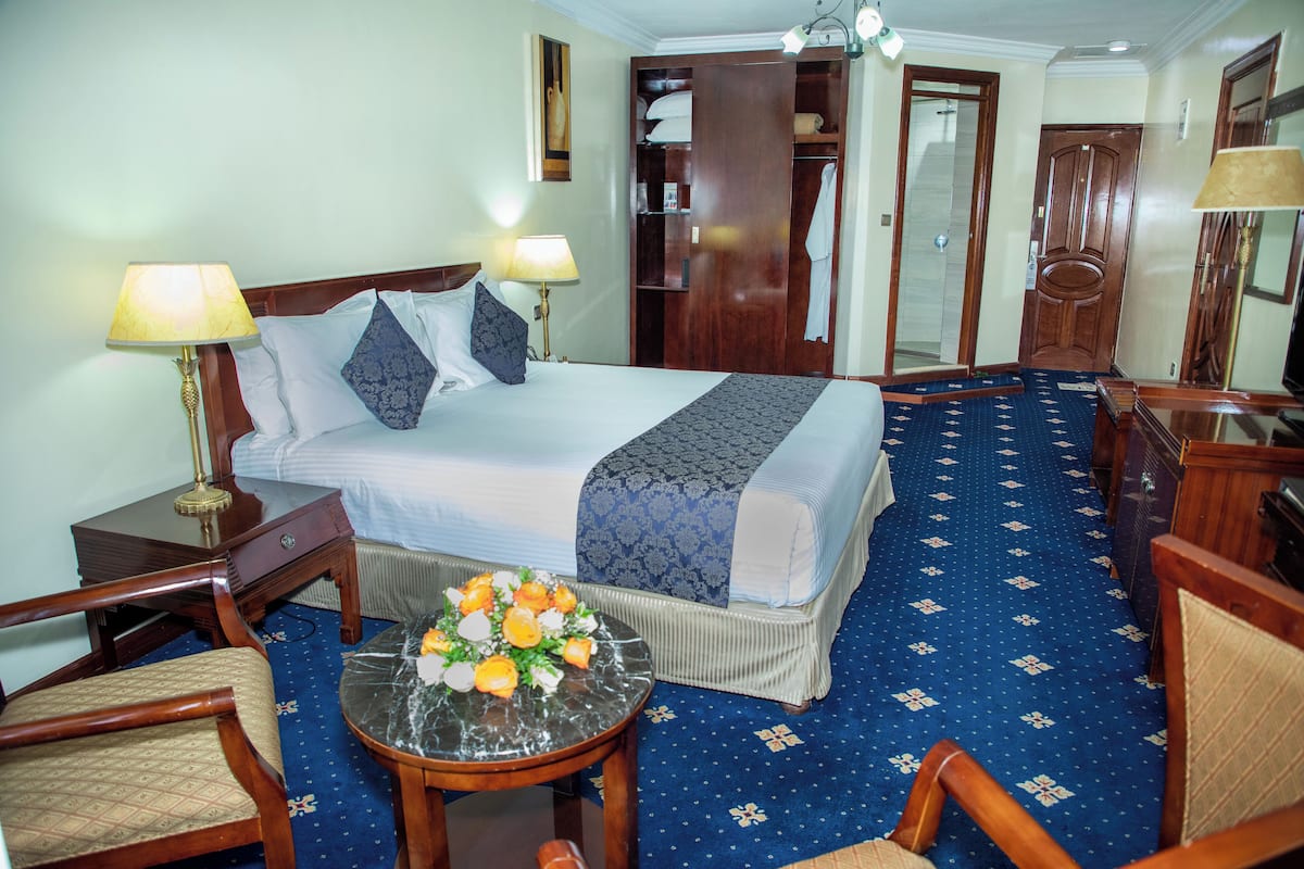 Una habitación de hotel con cama, dos lámparas de noche, una mesita con un arreglo floral, una silla y un armario.