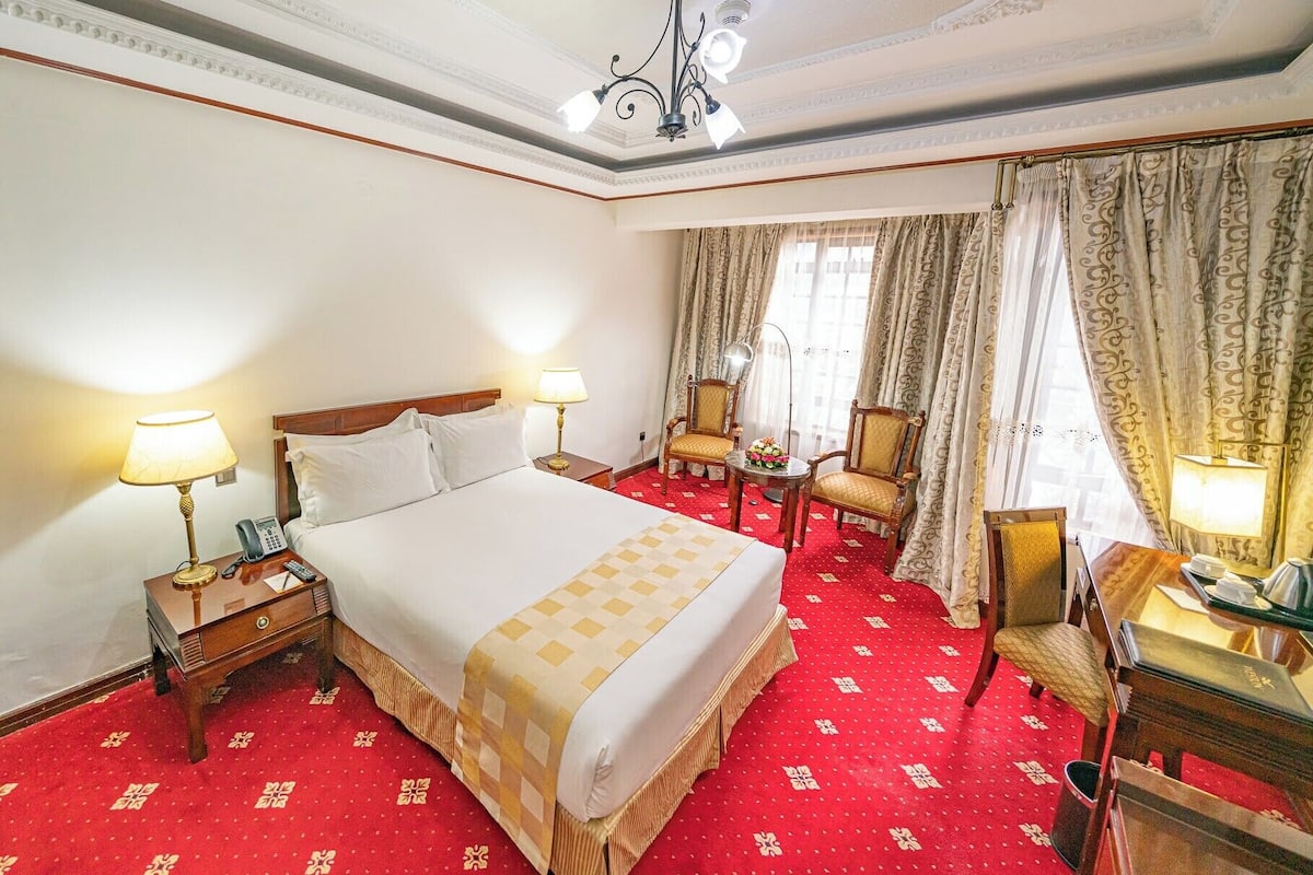 Habitación de hotel con una cama grande, dos mesitas de noche con lámparas, un escritorio con silla y un ventanal con cortinas.