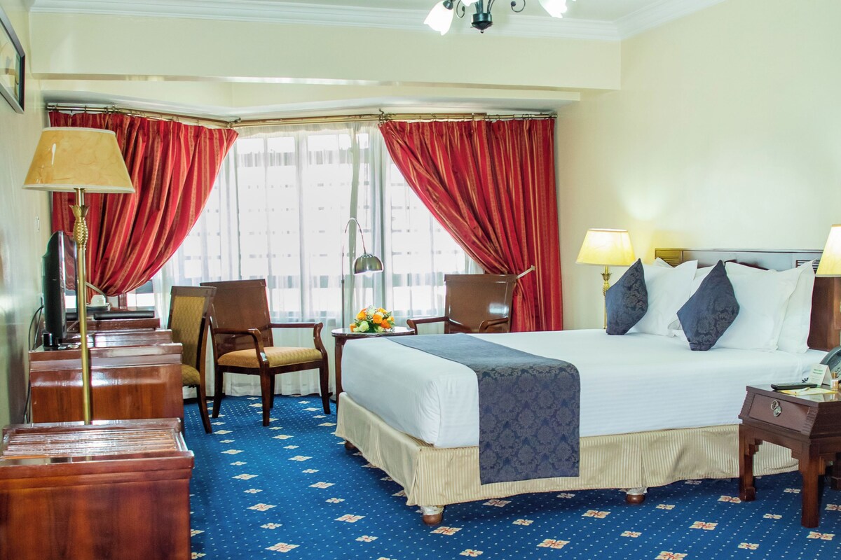 A hotel room with a bed, two chairs, a desk, a lamp, and a window with curtains.