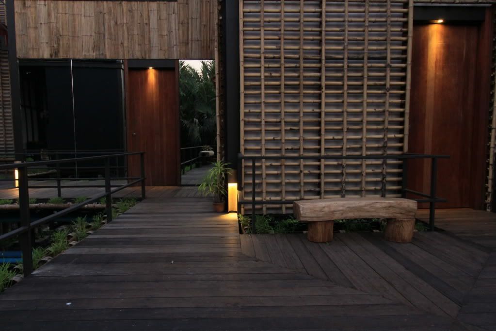 A wooden deck with a bench and railing, leading to a building with a bamboo wall and a door.